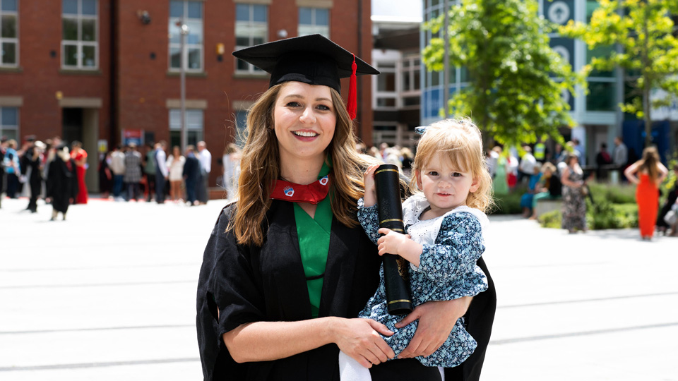 A smiling Emily wears her graduate gown and cap and is carrying her daughter, a toddler, who is holding the degree case. 