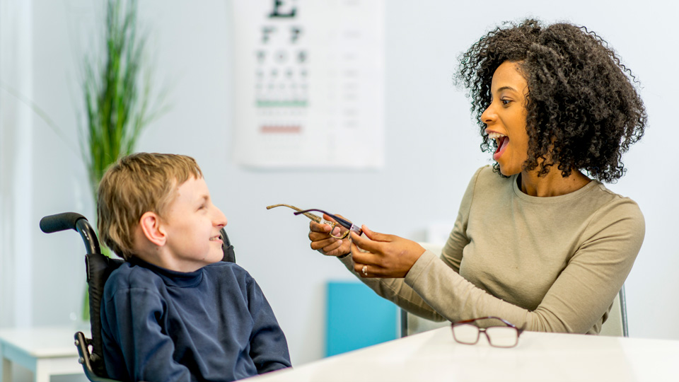 A young boy in a wheelchair smiles as an eye care practitioner shows him a pair of spectacles