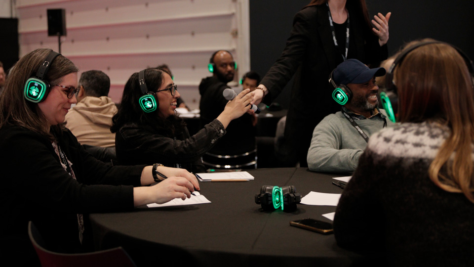 A handful of people are seated at a circle table in an event, each wearing glowing green earphones. A microphone is handed to one delegate
