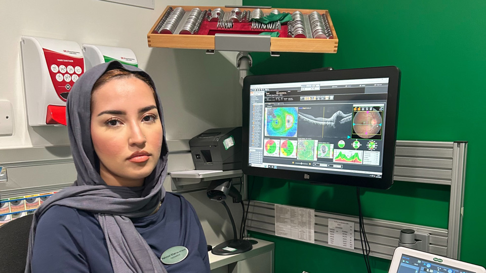 Mahnoor in a navy long-sleeved dress and headscarf sits at her computer in the testing room. Behind her the screen displays scan results