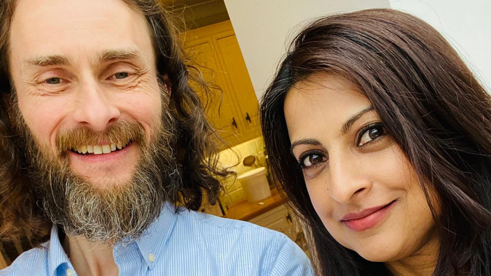 In a blue striped shirt, Ed, stands smiling with Riz, in a patterned floral blouse