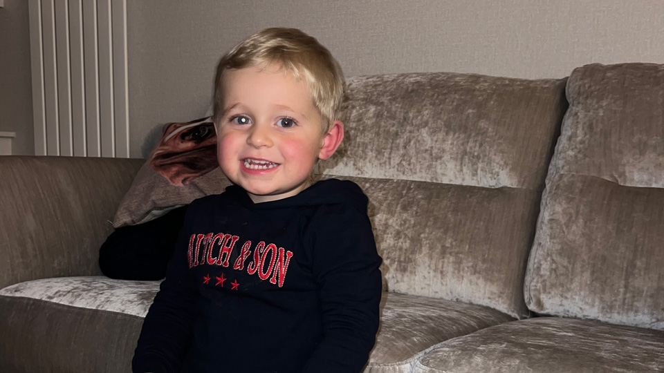 A toddler leans against a sofa holding a toy truck. One of his eyes is glowing with a white circle
