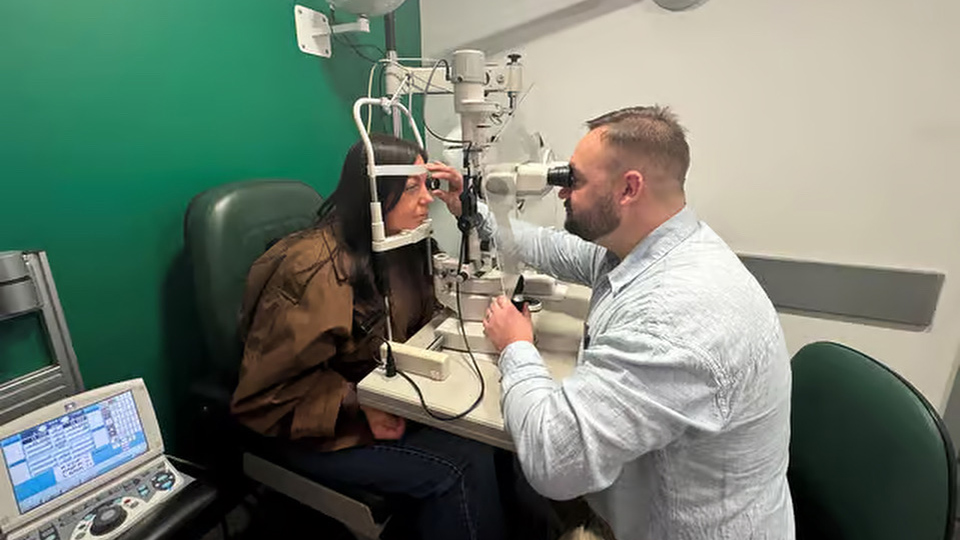 An optometrist, Craig, is carrying out an eye test on a patient using a slit lamp and a Volk lens. The walls are green, indicating this is a Specsavers