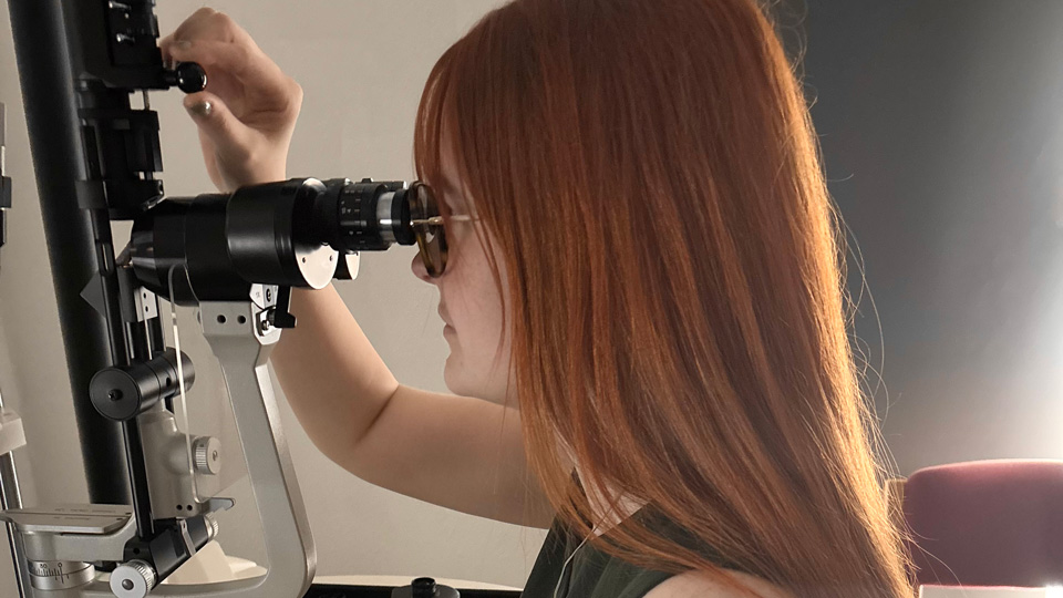 Lucie is practising with a slit lamp at university, her eye against the viewfinders while she controls it with a joystick and adjusts with her other hand
