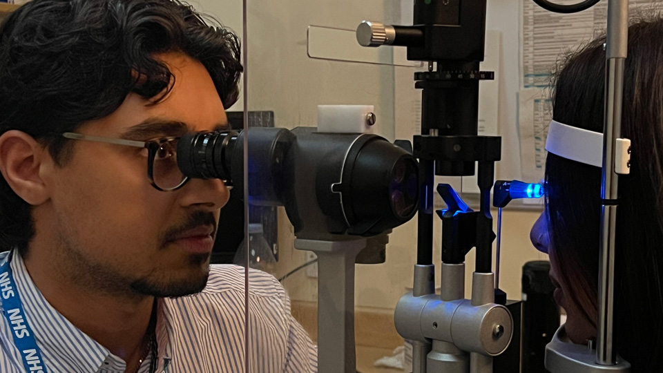 An optometrist wearing an NHS lanyard, Zak Saleem, examines a patient’s eyes using a slit lamp microscope in a clinical setting