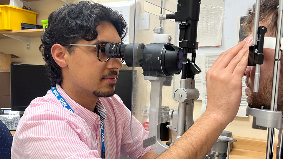 An optometrist wearing an NHS lanyard, Zak Saleem, examines a patient’s eyes using a slit lamp microscope in a clinical setting, with a blue light shining into the patient’s eye as she rests her chin on the support