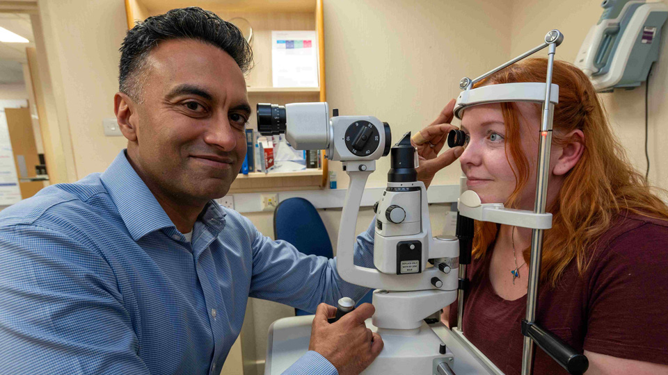 Sameer and Hannah pose during a sight test. Sameer has turned to the camera. With one hand he holds the control for a slit lamp, and with the other he holds a volk lens up to Hannah’s eye