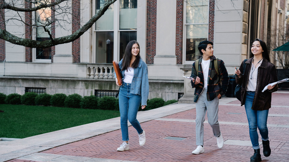 Three students walk through campus carrying paperwork and folders