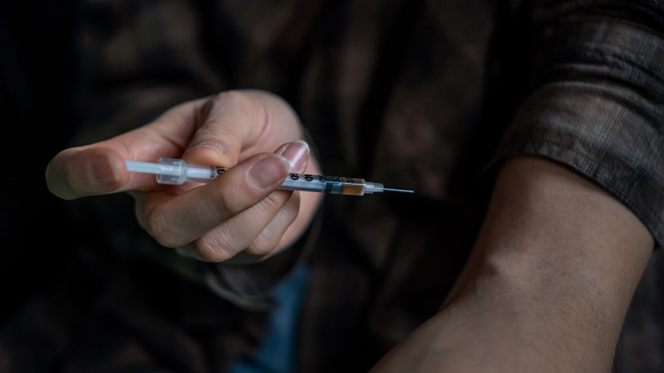 A man is shown with a hypodermic needle preparing to inject his arm
