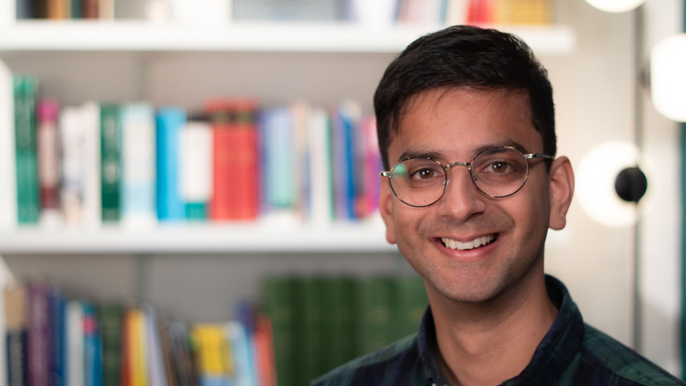 Karan is photographed in the library of the AOP offices, with rows of optometry and professional books behind him. He wears thin metal glasses and a checkered blue and green shirt