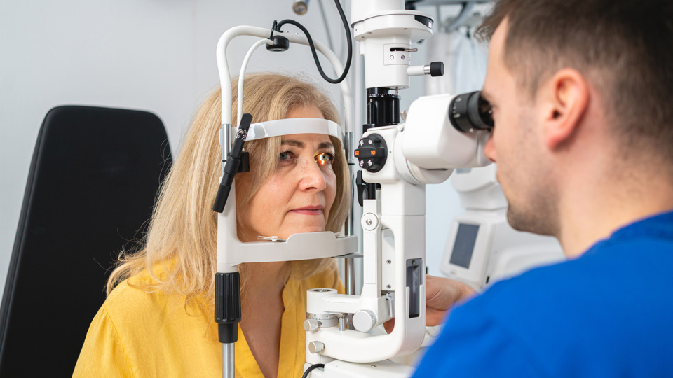 A woman in middle age has her chin on the rest of a slit lamp device for an eye test. On the right-hand side, the optometrist in blue scrubs is controlling the device to perform the test