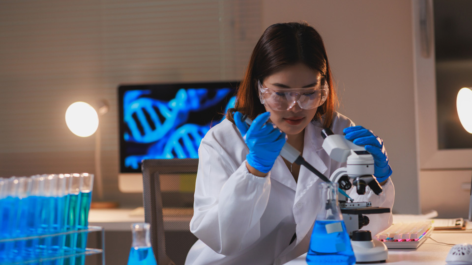 A scientist looks into a microscope with an image of a DNA double helix on a screen in the background