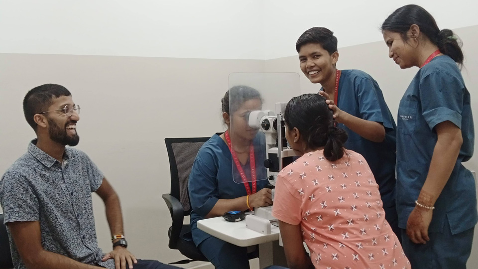 Benji sits to the left observing medical students practising an eye test. Three students are gathered around the slit lamp with a volunteer patient