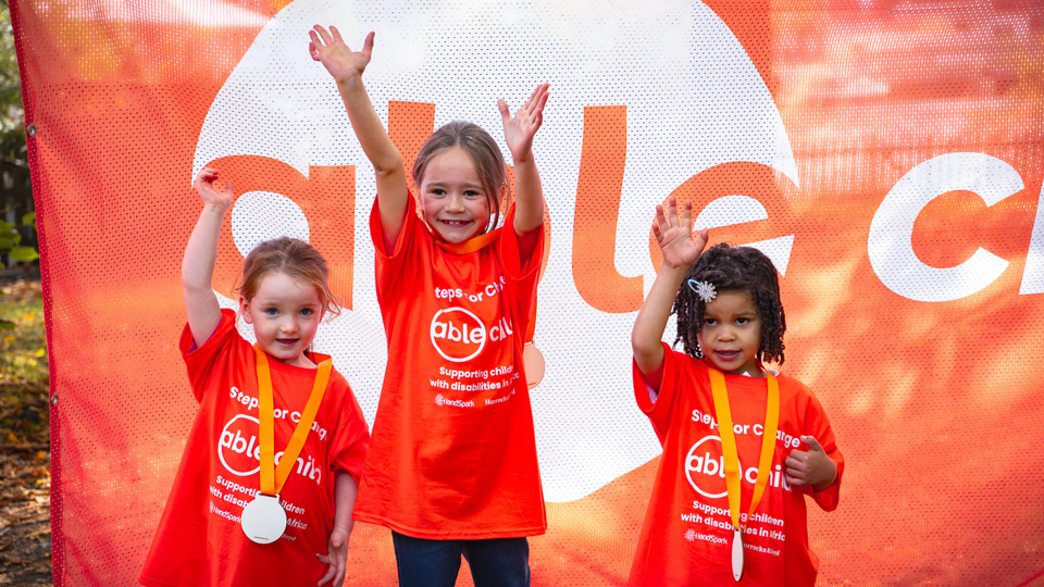 Three children stand in front of a large orange and white Able Child banner each wearing orange Able Child challenge t-shirts and medals. They are cheering for the camera