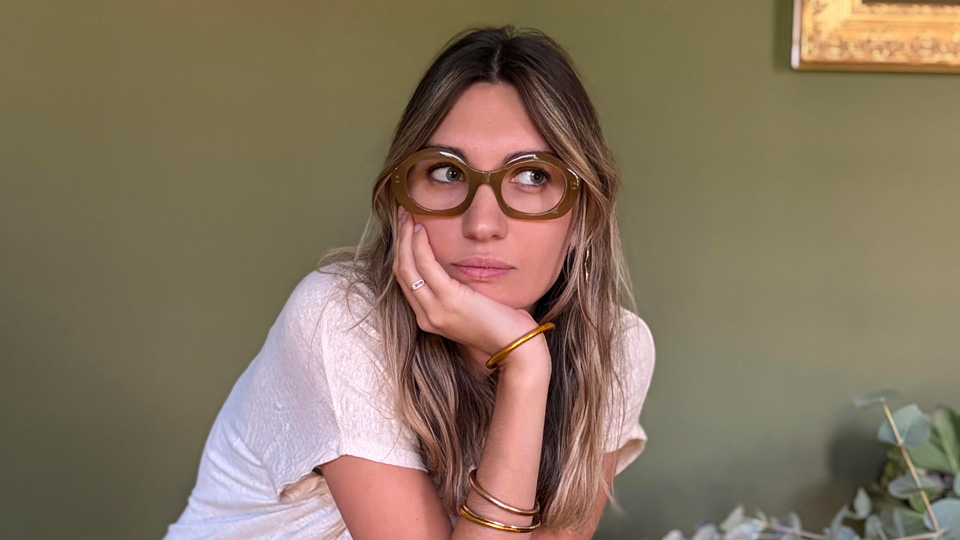 A young woman sits on a bright pink stool in front of an olive-green wall. She wears oversized oval spectacles in a deep brown, a white t-shirt and green trousers. She rests her chin on her hand and holds another pair of frames in her other
