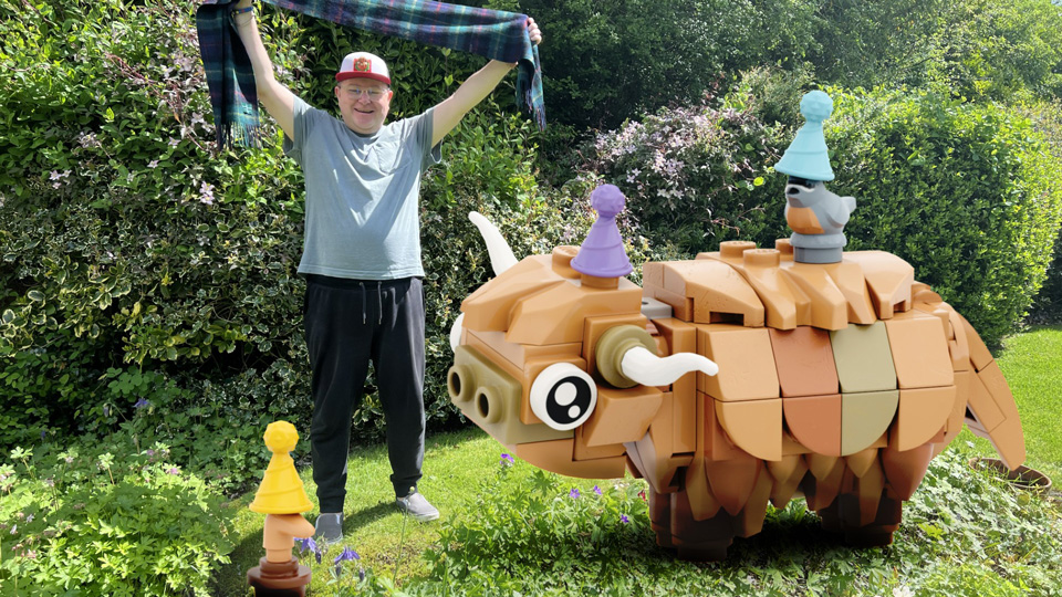 A man stands in a sunny garden holding a tartan scarf above his head beside a large orange, turtle-like garden sculpture with cartoon eyes, horns and pointed legs. Smaller colourful sculpted figures stand nearby among the flowers and greenery