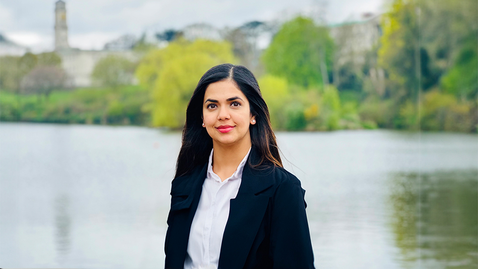 Roma is stood outside wearing a black suit jacket, in front of a lake