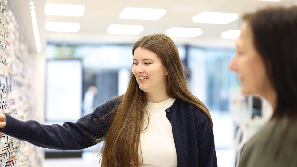 A young woman in an optician’s shop smiles as she reaches toward a wall display of glasses frames, while another woman stands slightly out of focus nearby, watching. Bright overhead lighting and a clean, modern interior create a welcoming retail environment