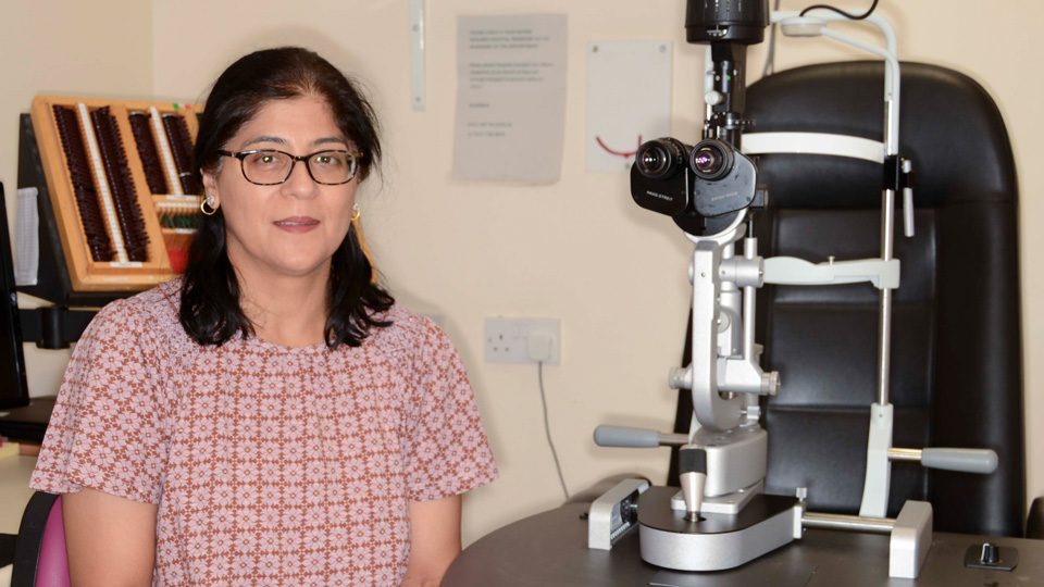 A woman with shoulder length dark brown hair and a red and white top checked with flowers is sitting to the left of a large piece of optical equipment and looking at the camera