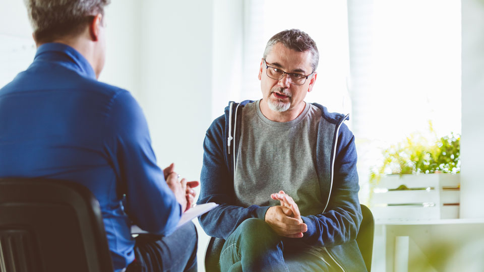Mature man talking with another man who is wearing a blue shirt and has his back to the camera. They are both sitting down