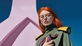 A young woman with bright orange hair stands in harsh sunlight, looking down towards the camera. She wears large butterfly-shaped frames with three contrasting colours of acetate on the top corners. She holds a pink striped snake against her chest. The general effect is striking, and a little science-fiction