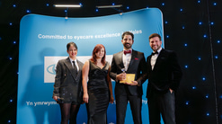 A group of four people dressed in formal evening wear stand in front of a blue event backdrop that reads “Committed to eyecare excellence in Wales.” One person holds a glass award and an envelope, suggesting a presentation or award ceremony. All four are smiling and posing for the photo under soft stage lighting, with small blue lights scattered across the dark background