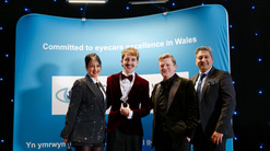 Four people dressed in formal attire stand smiling in front of a blue backdrop that reads “Committed to eyecare excellence in Wales.” The backdrop is set against a dark background lit with small blue lights. From left to right: a woman in a sparkly suit holding a microphone, a person in a dark red velvet jacket and bow tie, and two men in dark suits. All pose together on a stage under bright event lighting