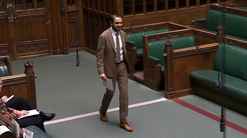 A man in a suit walks across the floor of the House of Commons chamber holding a sheet of paper, smiling slightly, with green leather benches, wooden panelling and hanging microphones visible around him