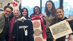 A group of six people pose inside, One holds a ‘Healthy eyes save lives’ poster, one holds a colour vision test, and another holds a sketch of Askew Eyewear