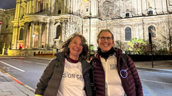 Two women in puffy coats and Vision Care t-shirts pose, smiling, in front of St Pauls Cathedral. The skies are dark and streetlights glow