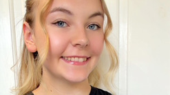 Lauren has long blonde hair, which curls round her face as she smiles at the camera. She wears a smart black t-shirt