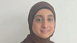 Saahirah sits side-on and smiles to the camera. She wears a chestnut brown headscarf and matching blazer