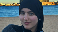 Sara is photographed on a beach, with a blue stretch of water reflecting the blue skies, and building lit up in the light of a setting sun beyond. Sara wears a black headscarf and a dark grey cowl jumper, with headphones around her neck