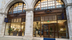 Boots Opticians Wigmore Street pictured from outside. The building has large pale stone archways, with decorated pillars and arches. Within each arch are huge windows, with ornate dark wood frames. Inside, the practice is lit with warm lights. Gold lettering on the windows indicates the Boots Opticians brand