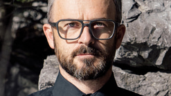 A man in grey square optical frames, wearing a black shirt and grey tie, is photographed in front of rocks. He has a deadpan expression