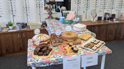 A table set up in an optometry practice, a sign hangs above it stating it is a coffee morning and a table is full of cakes