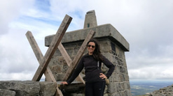 A woman wearing sunglasses and black hiking clothing stands beside a stone trig point at the top of a rocky mountain. She is smiling with one hand on her hip, leaning against a wooden stile built into a stone wall. Cloudy sky and distant landscape stretch out behind her