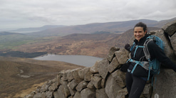 A woman wearing dark hiking clothes and a blue backpack leans against a stone wall on a mountainside. She is smiling toward the camera, with a wide landscape of hills, fields, and a lake stretching out below under an overcast sky.