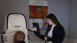A woman operates an eye-imaging machine while an older man sits with his face positioned in the device. She holds a control and looks at a monitor attached to the machine. A poster showing a retinal image is displayed on the wall behind them  