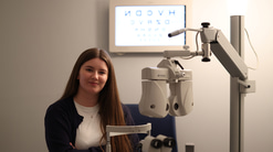 A woman stands in an eye-exam room with her arms folded, smiling softly. In front of her are pieces of optometry equipment, including a phoropter and a slit lamp, and an illuminated eye chart is visible on the wall behind her 