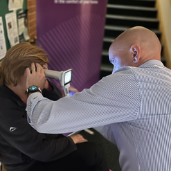 Matt uses a handheld imaging device to screen Linda’s eyes. In the background is a purple banner for OutsideClinic 