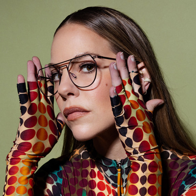 Eline touches her hands to either side of her thin metal aviator glasses. She wears a psychadelic dotty top with long sleeves that end as fingerless gloves
