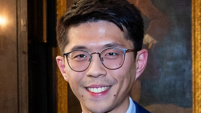 Fabian wears a navy suit and tire with white shirt. He has slim-line pantos style spectacles. He is photographed in a darkened room lit with warm yellow lights. A large painting of an approximately 17th or 18th century man