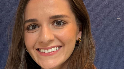 Yasmin has long curled brown hair past her shoulders and swept to the front. She wears a short-sleeved black top with white piping around the buttons. She is smiling