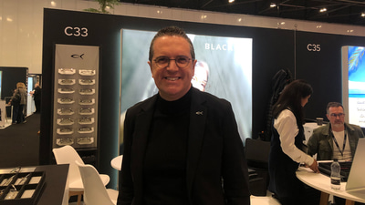 Nicola del Din is on the Blackfin stand at 100% Optical. He wears thin titanium glasses, a black turtleneck and a black blazer. On his lapel is a tiny metal fish – the Blackfin logo. Behind him, the stand design is sleek with black walls, titanium displays of shelves, and a light wall with model imagery