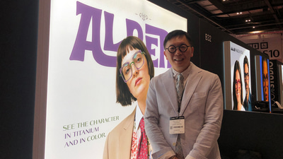 Diego Kim wears a pale striped suit and round circle glasses. He stands before a large light wall with the Albert I’mStein branding and a photograph of a model wearing a pair of lavender square spectacles