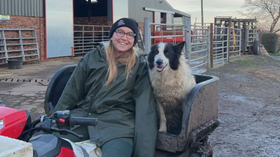 Holly sits in a quad bike wearing a green parka, wellies, and a beanie, next to hear is a sheepdog. In the background a barn is visible