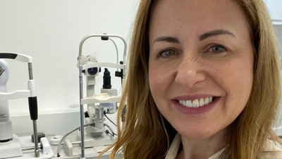 Lesley, in a white blouse, poses in the testing room at her practice. In the background can be seen optical equipment
