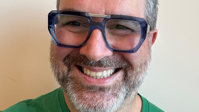 Peter smiles in a green t-shirt and navy blue Kirk and Kirk frames in an aviator style