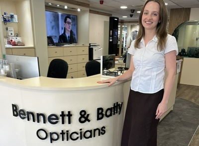 Charlotte stands at the reception desk for Bennett & Batty Opticians, wearing a long brown skirt and white blouse